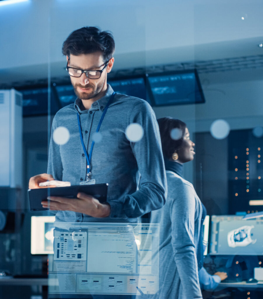 In Technology Research Facility: Chief Engineer Stands in the Middle of the Lab and Uses Tablet Computer. Team of Industrial Engineers, Developers Work on Engine Design Use Digital Whiteboard and Computers