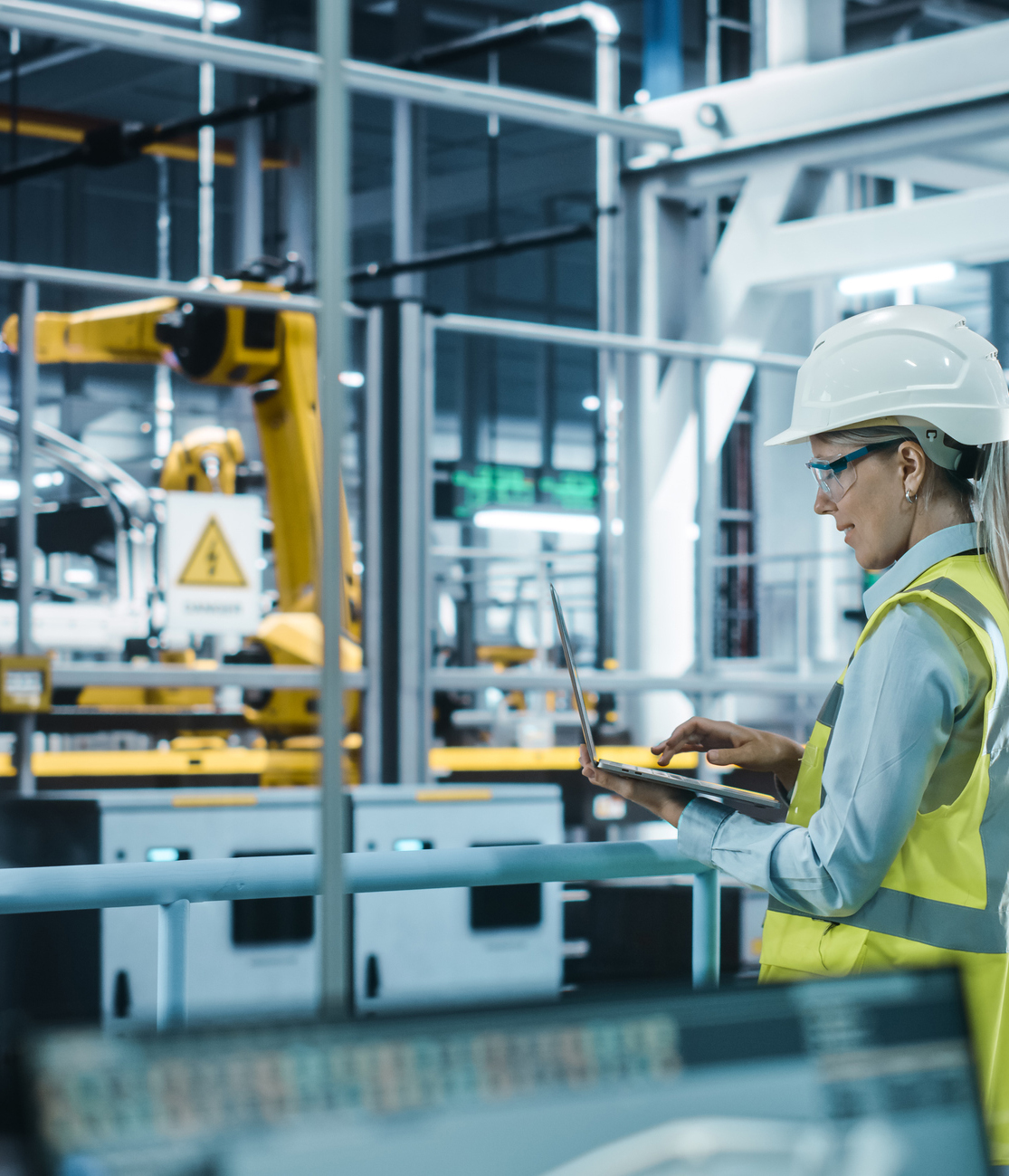 Car Factory: Female Automotive Engineer Wearing Hard Hat, Standing, Using Laptop. Monitoring, Control, Equipment Production. Automated Robot Arm Assembly Line Manufacturing Electric Vehicles. Car Factory: Female Automotive Engineer Wearing Hard Hat, Standing, Using Laptop. Monitoring, Control, Equipment Production. Automated Robot Arm Assembly Line Manufacturing Electric Vehicles.