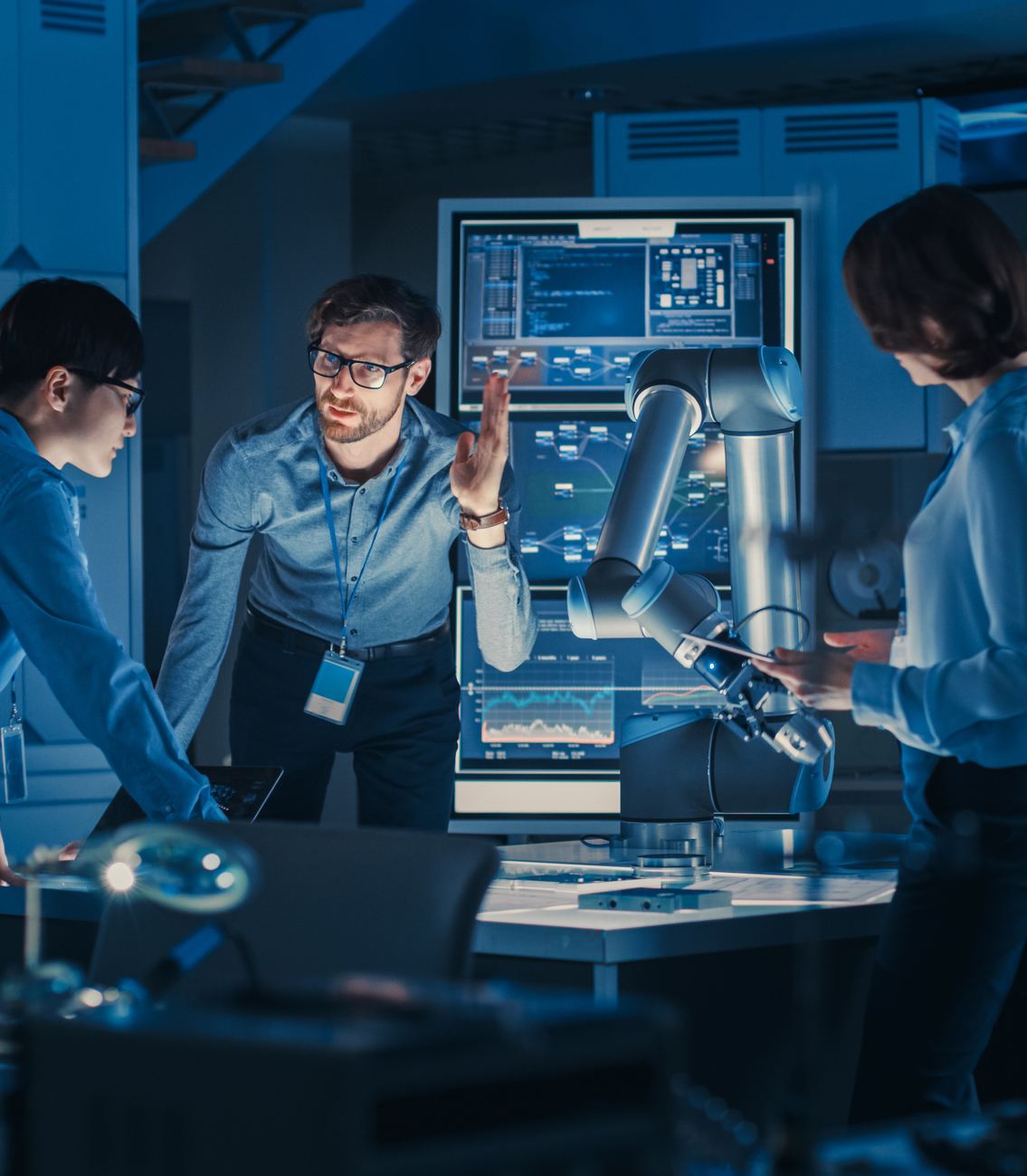 Diverse Team of Engineers with Laptop and a Tablet Analyse and Discuss How a Futuristic Robotic Arm Works and Moves a Metal Object. They are in a High Tech Research Laboratory with Modern Equipment.