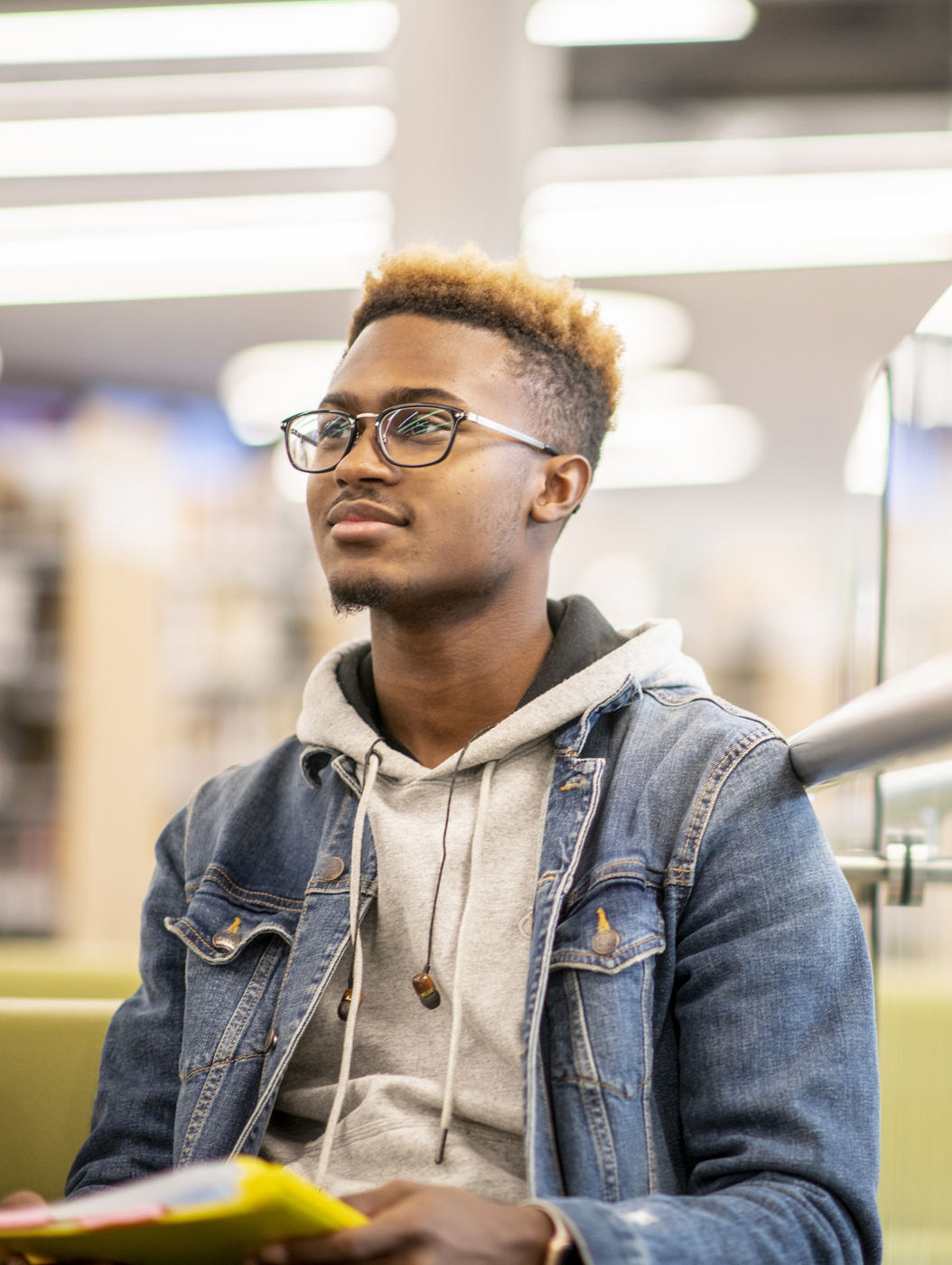 An African American university student is sitting in the library with his textbooks open on his lap, and studying